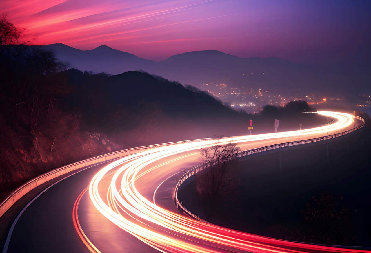 A winding road with bright red and white light trails from vehicles creates a dynamic curve, set against a twilight sky with deep pink and purple hues. Mountains are visible in the distance, and city lights illuminate the horizon.