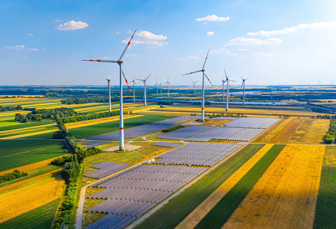 Wind turbines and solar panels in a green field on a sunny day. A large area with numerous wind turbines and solar panels under a clear sky. Fields stretch in different colours.