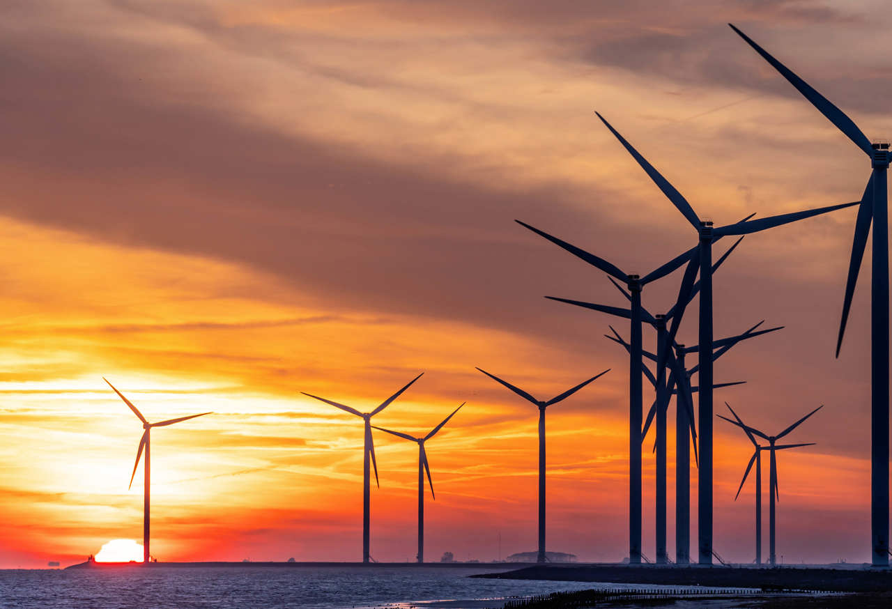 A series of wind turbines silhouetted against a vibrant sunset sky over a body of water. The sun is partially visible near the horizon, casting an orange and yellow glow across the scene.